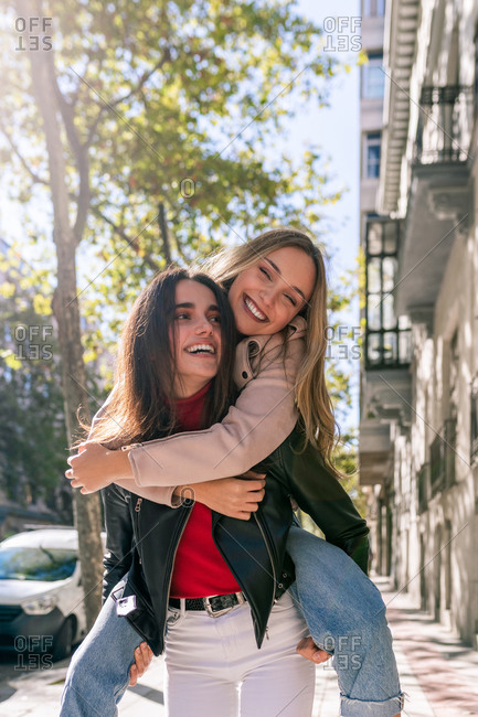 Positive woman piggybacking best friend while having fun on street and spending weekend together