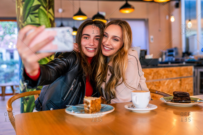 Satisfied female best friends sitting at table in cafeteria and taking selfie on smartphone while hugging and enjoying time together