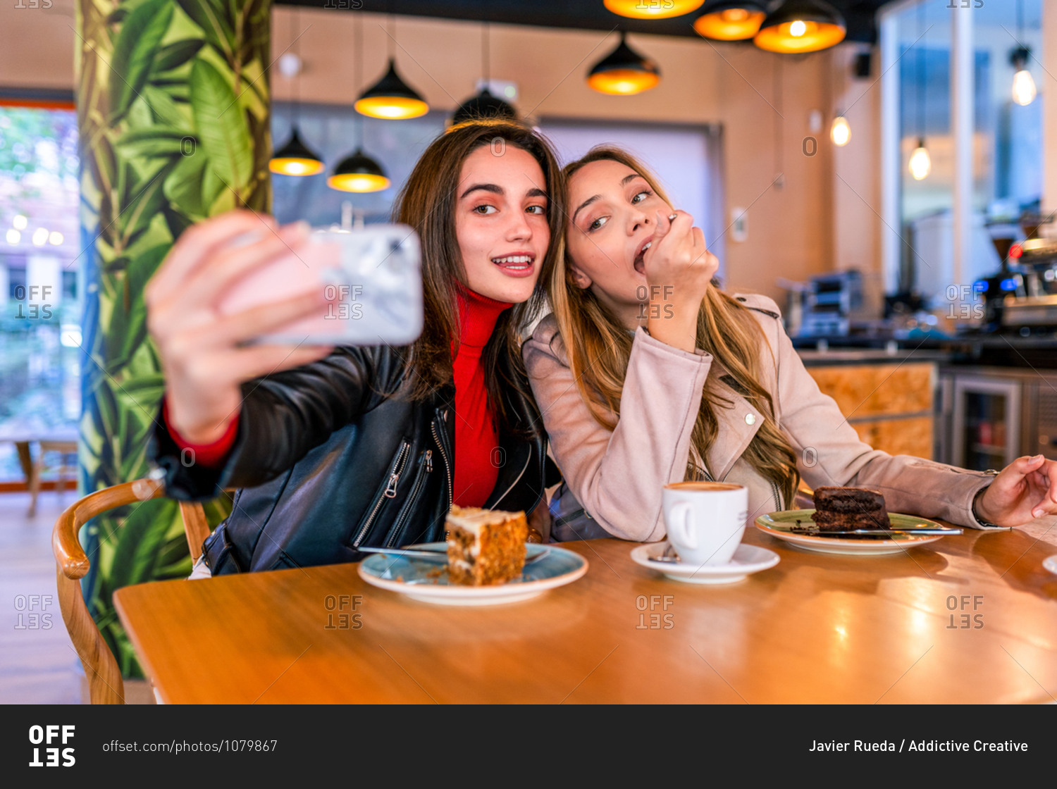Satisfied female best friends sitting at table in cafeteria and taking selfie on smartphone while hugging and enjoying time together