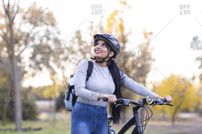Full body of positive young female in activewear and helmet riding bicycle through autumn forest with golden foliage in sunny day in countryside