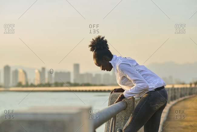 Side view of happy laughing young African American female in casual wear leaning over fence while enjoying free time on urban seafront