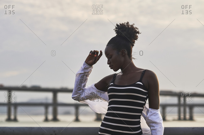 Tranquil pensive young African American female in casual clothes relaxing on urban seafront at sunset time