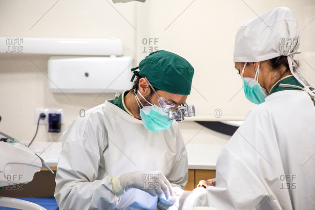 Focused professional dentist examining teeth of patent with dental instruments with help of assistant in bright room in clinic