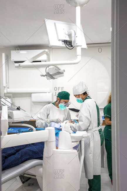 Dentists in masks and uniform operating patient lying in dental chair in bright modern clinic