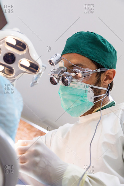 Low angle of focused male dentist in binocular glasses and uniform working in modern dental clinic