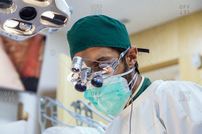 Low angle of focused male dentist in binocular glasses and uniform working in modern dental clinic