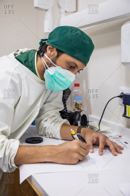 Male doctor in uniform and protective mask standing at table in medical room and writing notes in report