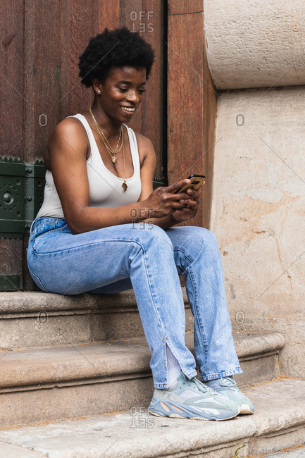 Cheerful curly haired African American female browsing social media in mobile phone and smiling brightly