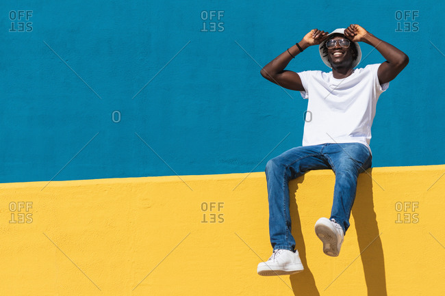 Low angle full body of optimistic young black male in casual outfit with panama hat and sunglasses smiling happily while siting on bright colorful fence and enjoying sunny summer day