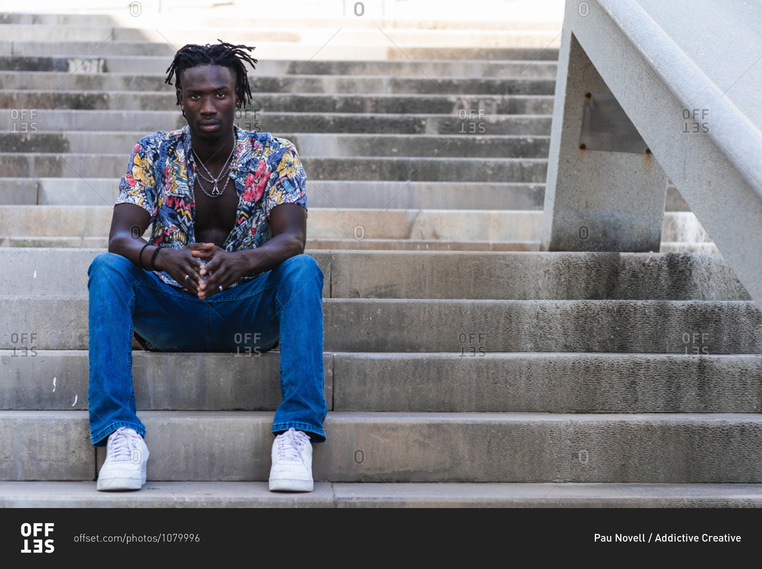 Low angle full body of serious African American male in colorful shirt and jeans with sneakers sitting on stone steps and looking at camera on urban street