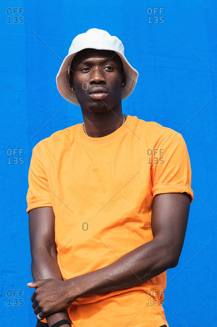 Young African American male in bright yellow t shirt and sportive pants posing while standing against blue wall
