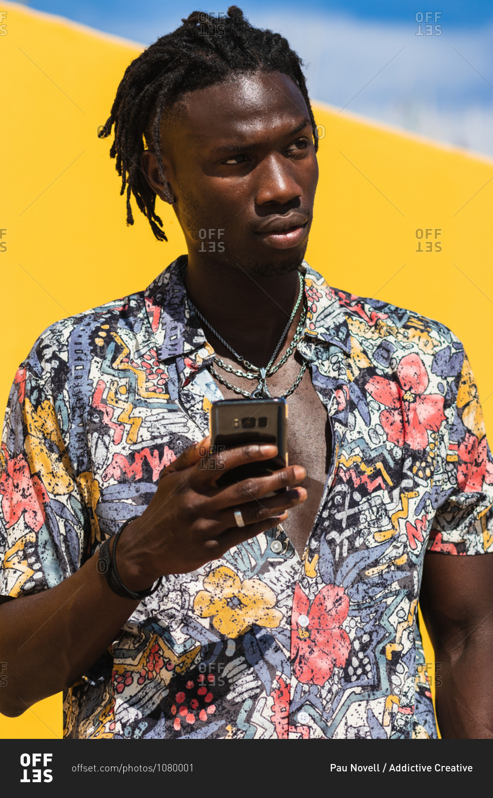 Confident young hipster African American male with dreadlocks wearing trendy summer shirt with floral print and jeans using mobile phone and looking way against yellow background