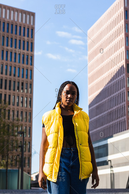 Low angle of young African American female in stylish yellow waistcoat standing against high rise buildings and cloudy blue sky on city street