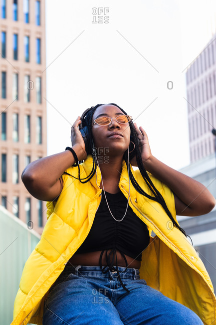 Low angle of young black female in trendy waistcoat and sunglasses touching headphones and enjoying favorite music with closed eyes in downtown