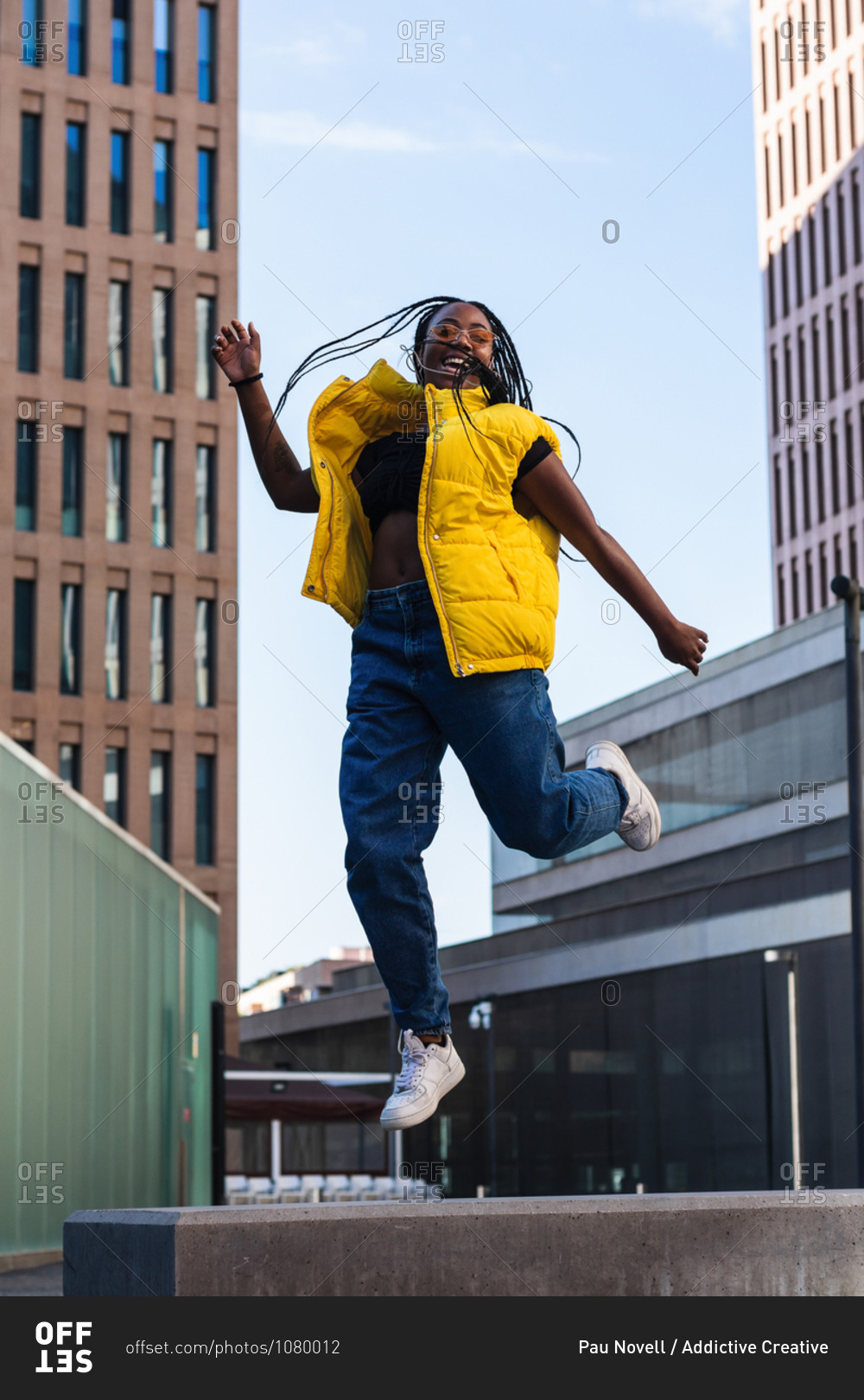 Happy black woman in trendy clothes laughing and leaping up on street of modern city looking at camera