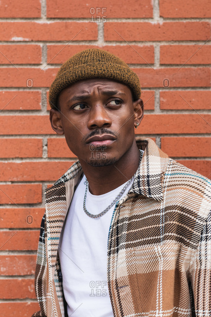 Young African American guy in checkered shirt and trendy hat frowning and looking away while standing against brick wall on city street