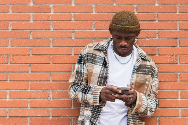 African American guy in casual outfit frowning and browsing social media on smartphone while leaning on brick wall on street