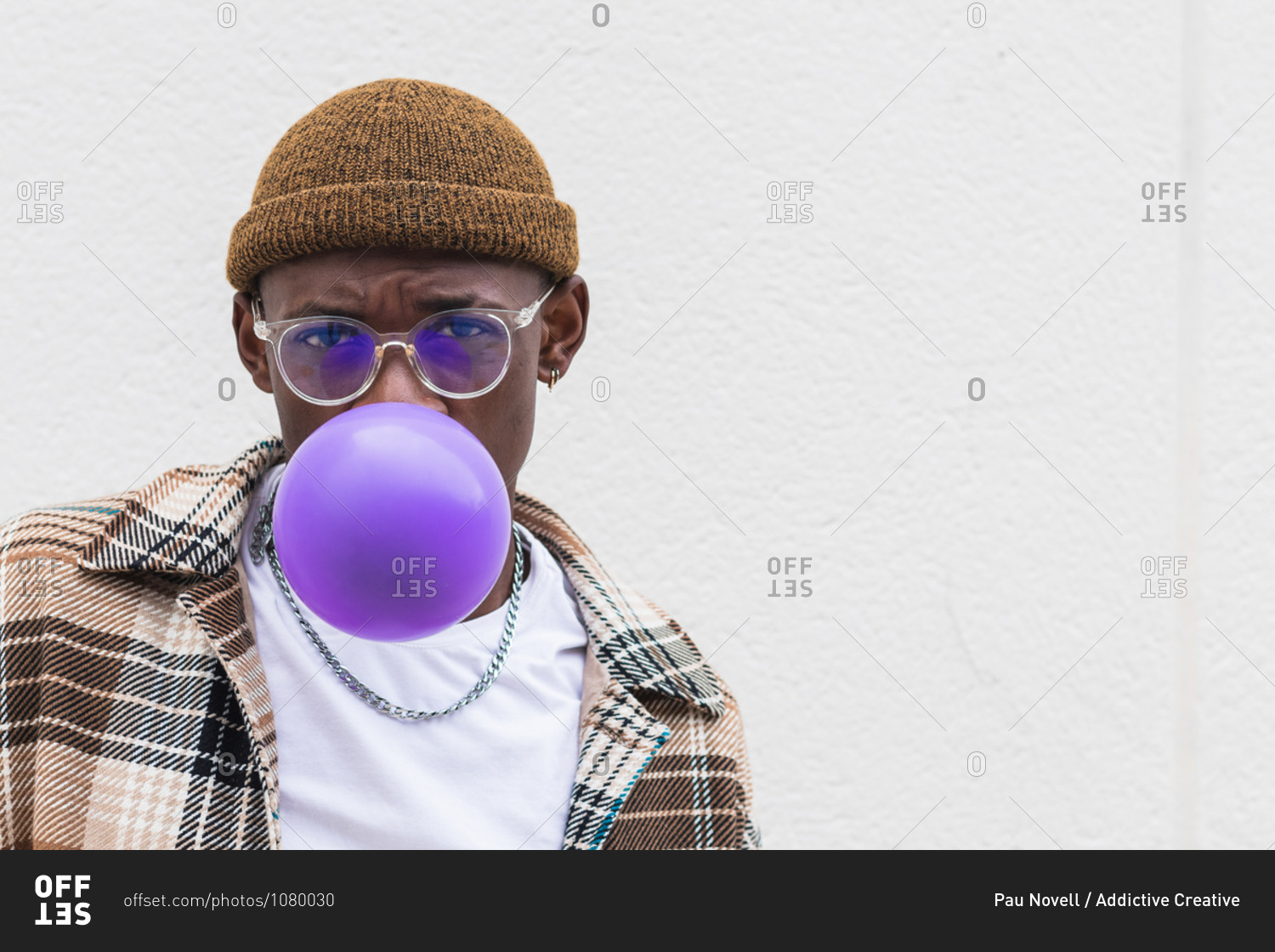 Young black male in stylish hat and glasses looking at camera and blowing violet balloon against gray wall