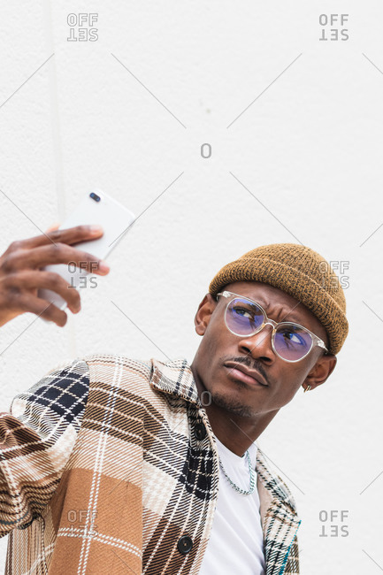 Trendy African American male in casual clothes and glasses frowning and taking selfie against gray wall on city street