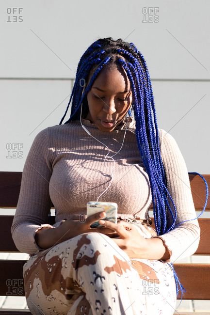 African American female with blue braids and in trendy outfit sitting on bench in city and chatting on cellphone while enjoying music in earphones