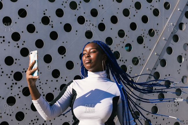 Determined African American female with blue braids standing in urban environment and taking photo on selfie camera of smartphone