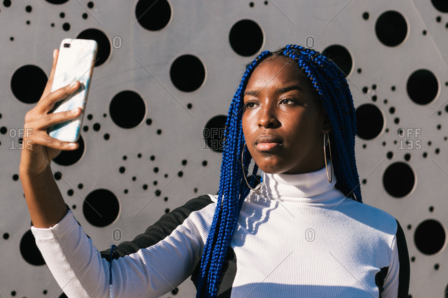 Determined African American female with blue braids standing in urban environment and taking photo on selfie camera of smartphone
