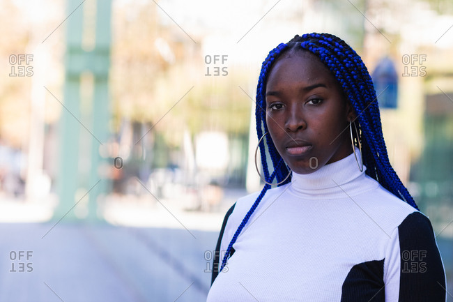 Calm African American female with blue braided hair standing in city and pensively looking at camera