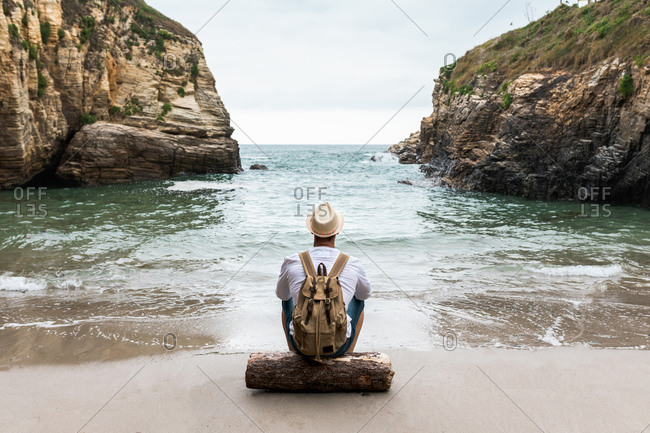 Back view of unrecognizable male backpacker in hat sitting on wooden log on coast near sea with rocky cliffs and relaxing during summer journey