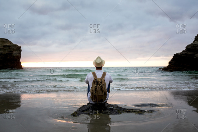 Back view of anonymous male traveler sitting on a rock at sandy coast enjoying views during summer vacation near sea at sunset