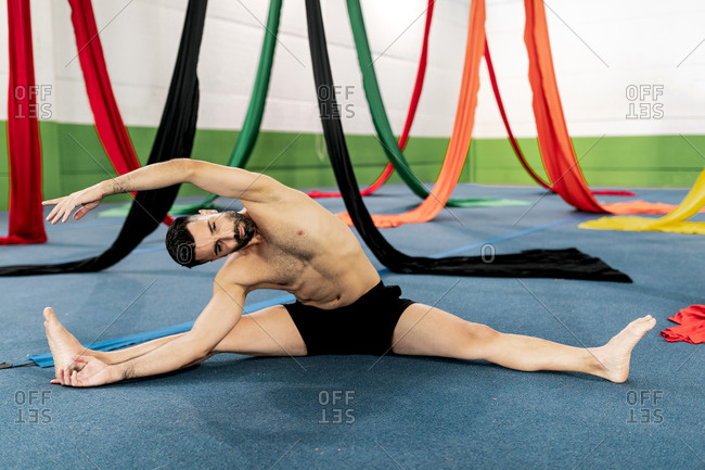 Barefoot muscular man sitting on floor near pieces of cloth and bending forward while stretching during aerial dance rehearsal in studio