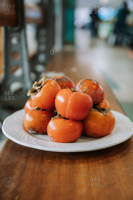 Pile of fresh ripe delicious persimmon fruits arranged on white plate placed on wooden counter in food market