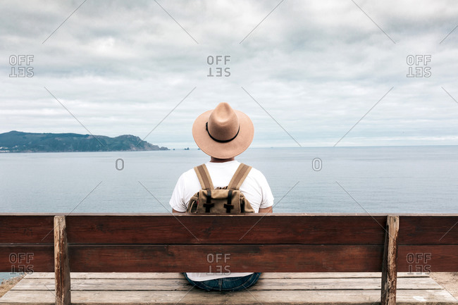 Back view high angle of male explorer in hat sitting on wooden bench and admiring spectacular scenery of sea and rocks