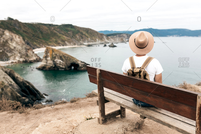 Back view high angle of male explorer in hat sitting on wooden bench and admiring spectacular scenery of sea and rocks