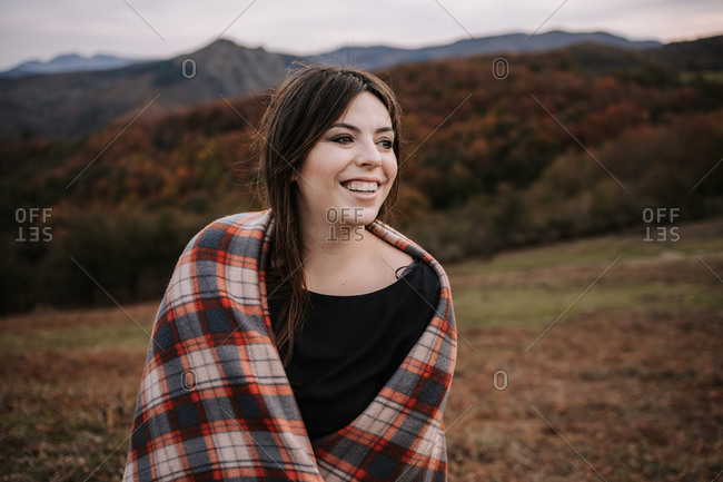 Delighted female wrapped in cozy checkered blanket standing on hill in highlands and looking away