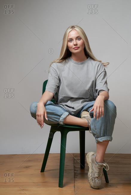 Young cool female in stylish outfit sitting on chair on gray background and looking at camera