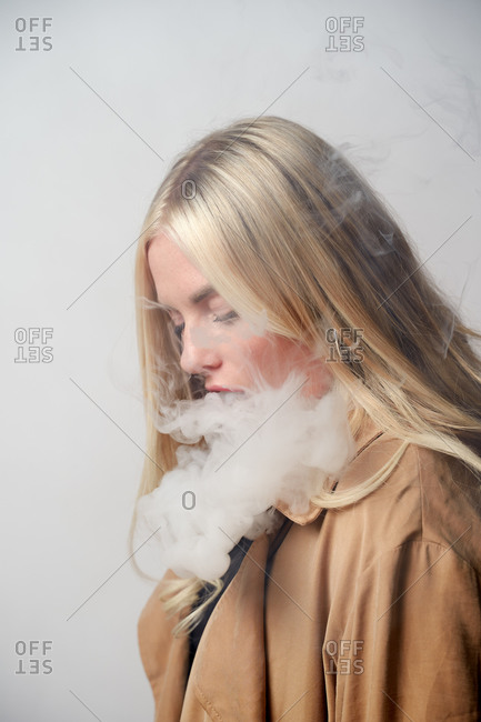 Tranquil female in stylish outfit standing in studio and smoking vape while looking down