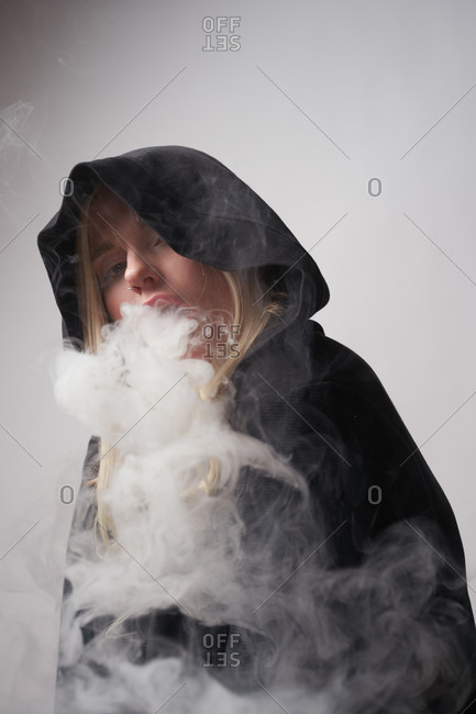 Rebellious young female in black hoodie smoking e cigarette and exhaling steam while looking at camera on dark gray background