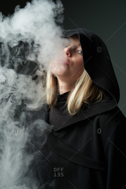 Rebellious young female in black hoodie smoking e cigarette and exhaling steam while looking at camera on dark gray background