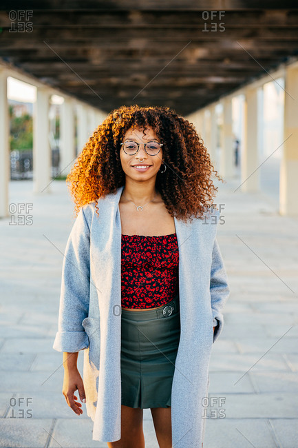Young black female in trendy outfit and glasses looking at camera while standing under roof on city street
