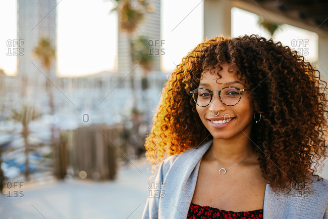 Young black female with curly hair in trendy outfit and glasses looking at camera while standing under roof on city street
