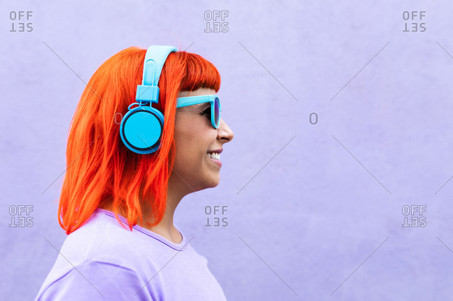 Happy stylish adult female with ginger dyed hair listening to music in headphones and singing along against violet wall in city