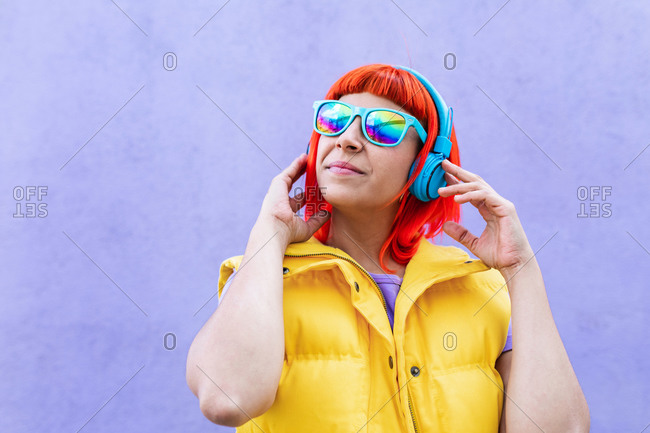 Happy stylish adult female with ginger dyed hair listening to music in headphones and singing along against violet wall in city