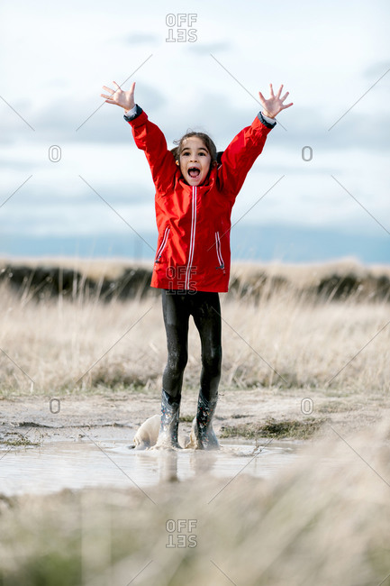 Carefree kid in raincoats and rubber boots splashing water in puddle while having fun and laughing on sunny day in countryside