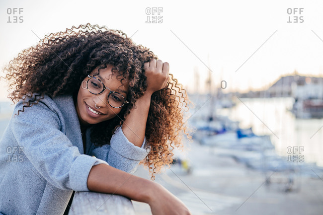 Positive ethnic female with curly hair smiling and looking away while leaning on fence in harbor