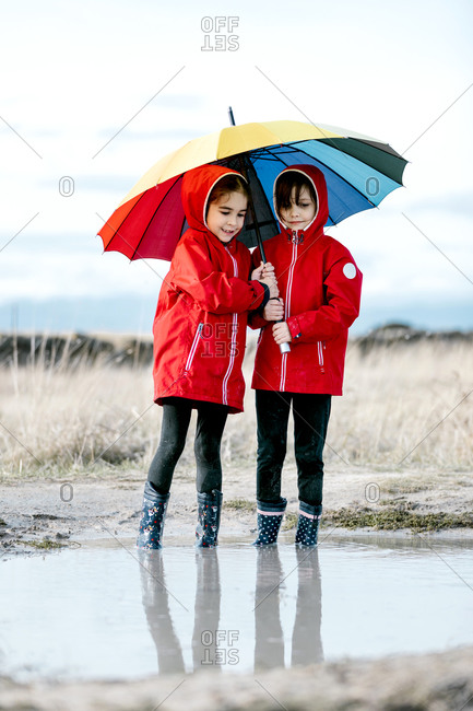 Excited sisters with colorful umbrella and in rubber boots playing in puddle while splashing water and having fun