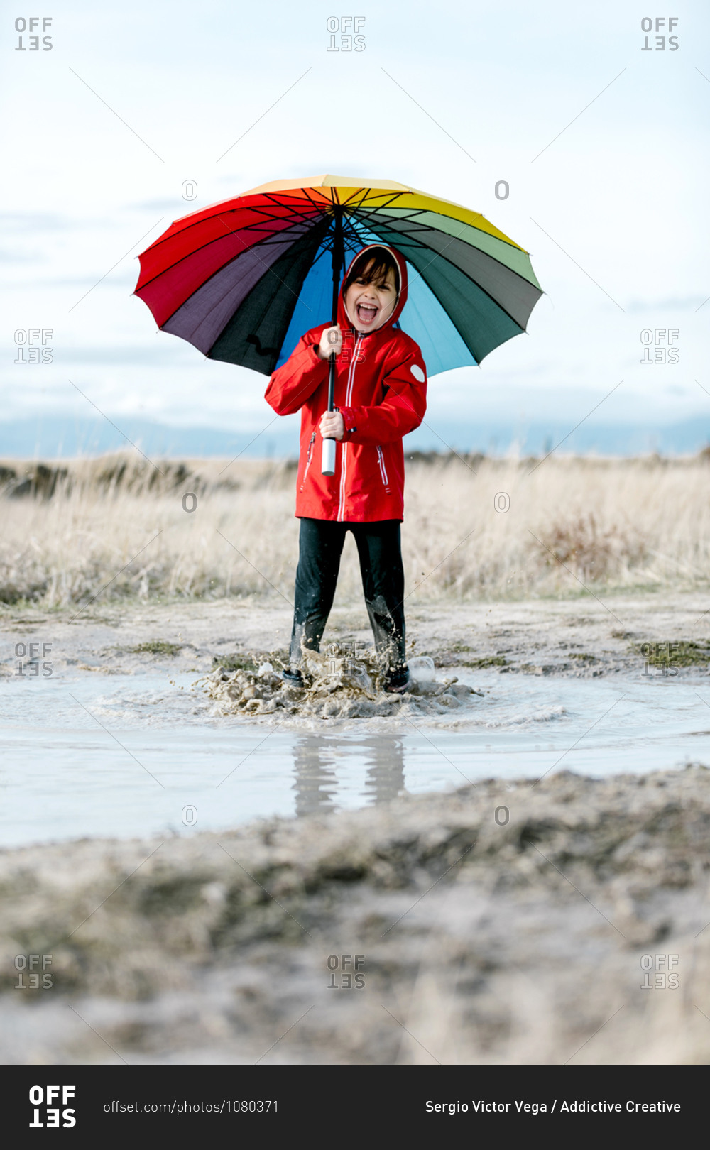 Excited little girl with colorful umbrella and in rubber boots playing in puddle while splashing water and having fun