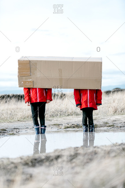 Anonymous children in rubber boots and raincoats standing in puddle and hiding behind cardboard package while having fun during weekend