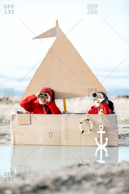 Playful children sitting in handmade cardboard boat in puddle and looking through spyglass while having fun at weekend together