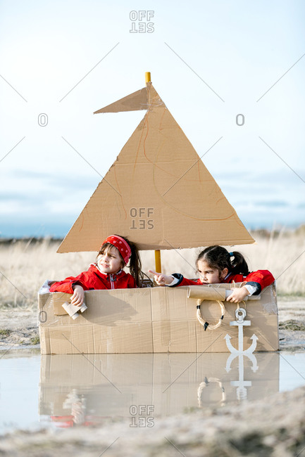 Playful children sitting in handmade cardboard boat in puddle and looking through spyglass while having fun at weekend together