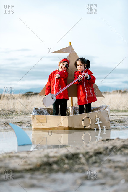 Optimistic children standing in carton boat with paddle above heads while having fun and enjoying game